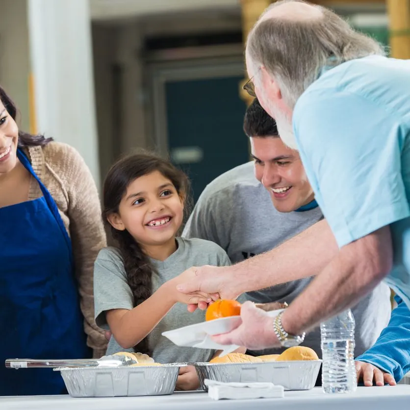 A volunteer handing a plate of food to a smiling girl at a community meal